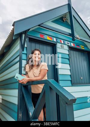 A woman standing on the steps of a colorful Brighton Bathing Box in Melbourne, Australia. The iconic beach huts are known for their vibrant stripes an Stock Photo