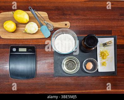 A top view of baking essentials on a wooden table, featuring lemons, flour, vanilla extract, oil, an egg, and lemon zest, alongside kitchen tools like Stock Photo