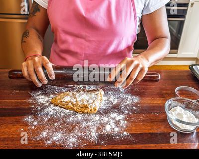 A woman in a pink apron uses a dark rolling pin to flatten dough on a floured wooden counter. A small glass bowl filled with flour is nearby, adding t Stock Photo