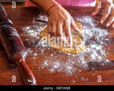 Hands using a circular cutter to shape cookie dough on a floured wooden surface. A rolling pin is next to it, indicating a baking process in a domesti Stock Photo