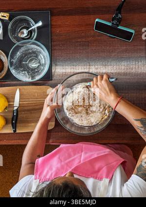 An overhead view of a woman mixing cookie dough in a glass bowl on a wooden countertop. Various kitchen utensils, ingredients, and a cutting board wit Stock Photo