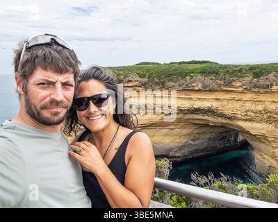 Two people smiles at the camera, enjoying a bright day by the ocean in the Twelve Apostles, Australia. They stand in front of a rugged cliff formation Stock Photo