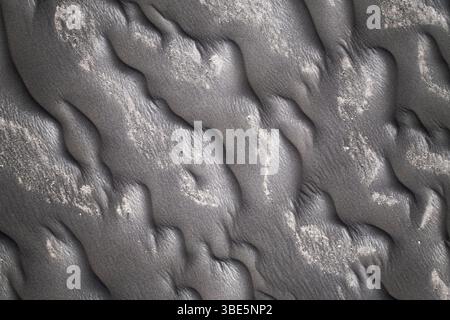 Close-up image capturing the intricate patterns formed on a black sand beach, showcasing textures and slight discolorations. Stock Photo