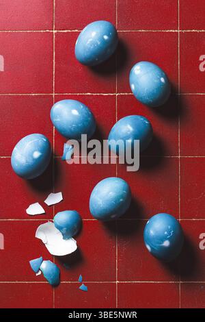 From above, a collection of blue Easter eggs on a red tiled floor, some whole and one broken with scattered pieces. Stock Photo