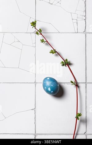 Top view of a single blue Easter egg adorned with a fresh green sprig on a textured white tiled background, symbolizing spring and renewal. Stock Photo