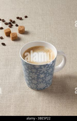 A warm cup of coffee with milk, resting on a beige fabric surface. The mug features a festive snowflake design, complemented by scattered coffee beans Stock Photo
