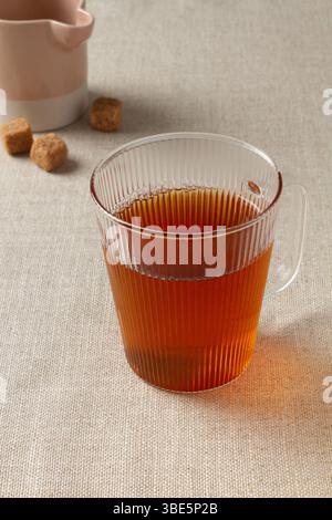 A warm cup of chai tea served in an elegant ribbed glass mug on a textured beige surface. Sugar cubes and a small jug are visible in the soft-focus ba Stock Photo