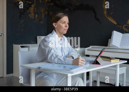 A woman sits at a table, preparing for a piano lesson in a music studio. She is taking notes and organizing materials, with sheet music and a tablet n Stock Photo