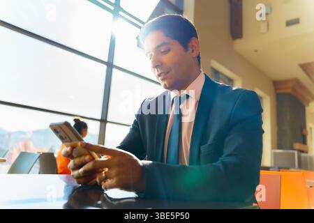 A young Chilean businessman in a suit concentrates on his smartphone while sitting in a bright, modern environment. Sunlight streams through large win Stock Photo