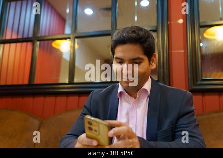 A young Chilean businessman in a suit, seated in a cozy cafe, engrossed in his smartphone. The warm lighting and stylish decor create a professional y Stock Photo