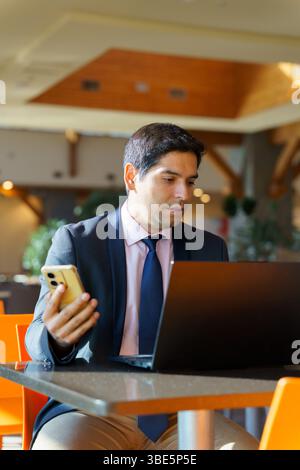 A young Chilean businessman in a professional setting, concentrating on his laptop while holding a smartphone. The modern setting background highlight Stock Photo