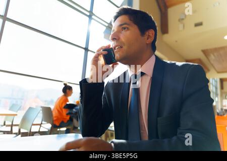 A young Chilean businessman, dressed in a suit, talks on the phone in a modern, sunny environment with large windows, concentrating on the conversatio Stock Photo