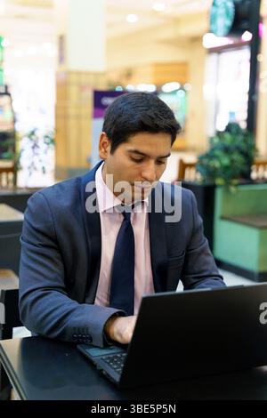 Focused young Chilean businessman wearing a dark suit and pink shirt, sitting at a desk in a modern cafe, concentrating on work on his laptop computer Stock Photo