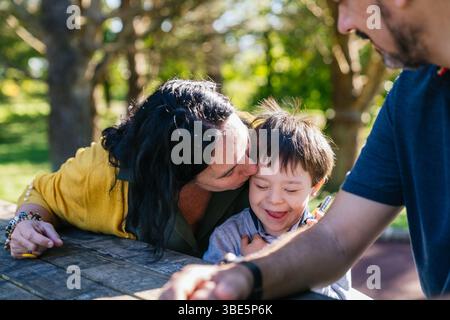 A heartwarming scene of a family enjoying a sunny day outdoors. A woman gently kisses a smiling child while sitting at a wooden table, surrounded by n Stock Photo