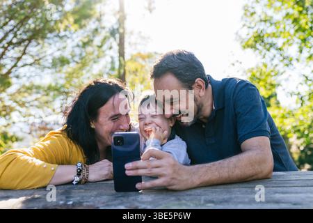 A cheerful family of three taking a selfie together outdoors on a sunny day. They are smiling and laughing, embodying happiness and closeness in a vib Stock Photo