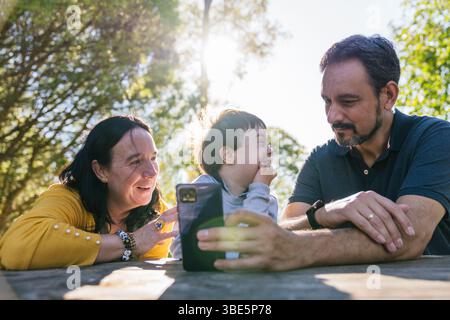 A family shares a happy moment in the sunshine, with a child with Down syndrome smiling between two adults. They gather around a phone, creating new m Stock Photo
