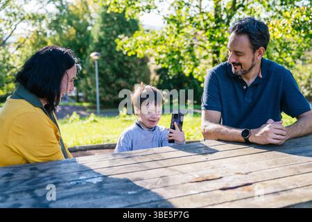 A joyful moment shared by a family at an outdoor picnic table. The parents watch their child with Down syndrome using a smartphone, surrounded by lush Stock Photo