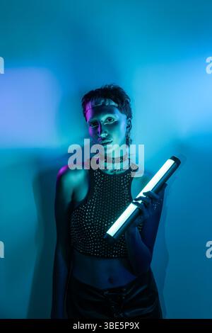 A striking cyberpunk-themed model poses under vibrant neon lights, holding a futuristic light stick. Her edgy style embodies a modern blend of technol Stock Photo