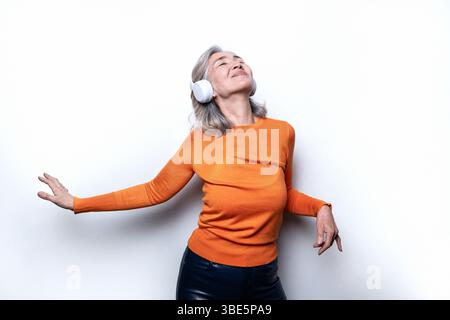 Asian Chinese senior woman wearing headphones and an orange sweater, enjoying music with a joyful expression, standing against a plain white backgroun Stock Photo