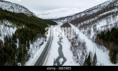 A breathtaking aerial view captures a Nordic winter landscape, showcasing a road alongside a partially frozen stream framed by snow-covered trees and Stock Photo