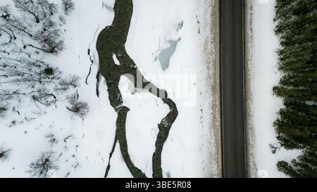 Aerial view of a Nordic landscape showcasing a road flanked by a frozen stream and snow-laden trees. The serene winter scene captures nature’s tranqui Stock Photo