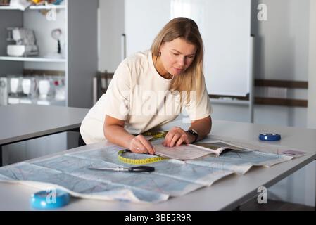 A woman is focused on her task in a sewing workshop, measuring fabric with a tape measure and working diligently. The room is filled with sewing equip Stock Photo