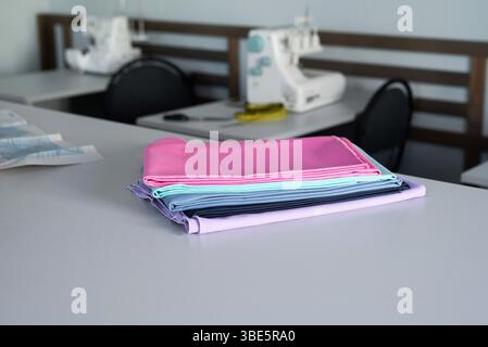 A stack of colorful fabrics on a table in a sewing workshop, with sewing machines and tools in the background. A woman is actively working on a projec Stock Photo