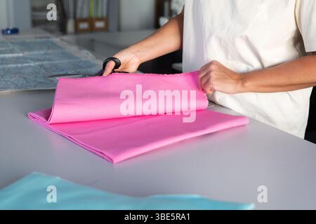 A woman carefully cuts a piece of pink fabric on a table in a sewing workshop. With focus and precision, she prepares materials for her next sewing pr Stock Photo