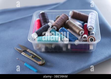 A clear plastic box filled with colorful thread spools sits on folded blue fabric next to small scissors and a seam ripper, showcasing a sewing worksh Stock Photo