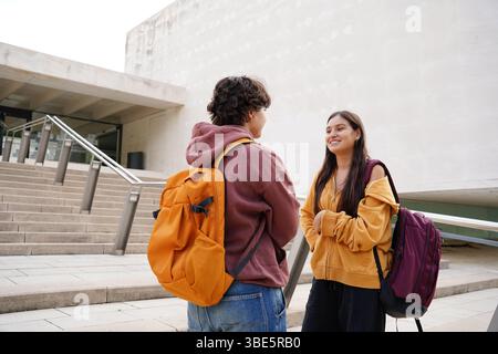 Happy non-binary teenager and a female teen engage in a friendly conversation while standing outside of their school. Both are dressed in casual attir Stock Photo