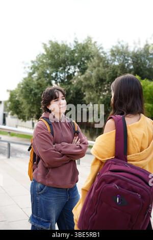 Two teenagers, one non-binary with short hair in a maroon hoodie and the other a female in a yellow cardigan, engaged in a friendly conversation while Stock Photo