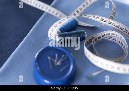A close-up view of sewing tools including a white measuring tape, two blue thread spools, and a pin holder placed on blue fabrics, representing a sewi Stock Photo