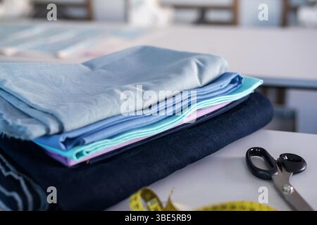A neatly folded stack of colorful fabrics rests on a table alongside tailoring scissors and a yellow measuring tape, representing sewing, fashion desi Stock Photo