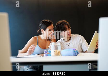 A non-binary teenager and a female student sit closely together at a school desk, engaged in shared educational activities. The non-binary individual Stock Photo