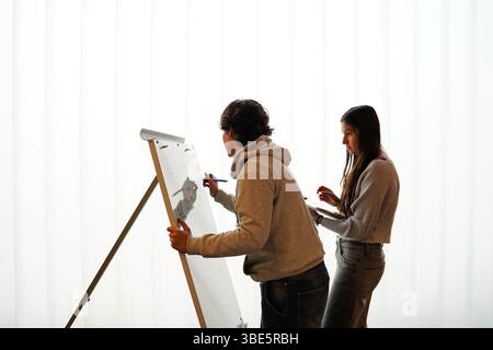 Two teenagers, a non-binary individual and a female, participate in an writing activity, o a whiteboard easel in a brightly lit classroom environment. Stock Photo