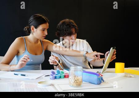 Female and non-binary teenager studying together, focused on a digital tablet and surrounded by artistic supplies. They engage in a collaborative and Stock Photo