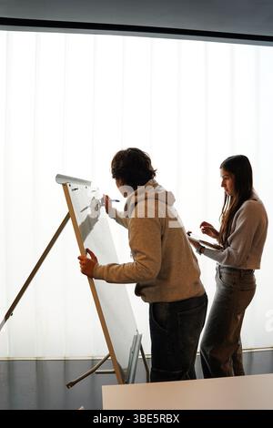 Two teenagers, a non-binary individual and a woman, collaborate on a writing project in a bright, modern classroom. They are focused on their artwork, Stock Photo