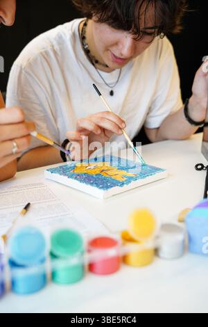 Two teenagers focus on painting during an art class at school. The non-binary individual paints a canvas while discussing techniques with a female com Stock Photo