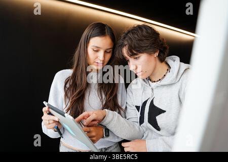 Two young students, a non-binary teenager with short hair and a female student with long hair, are engaging in scholastic activities using a digital t Stock Photo