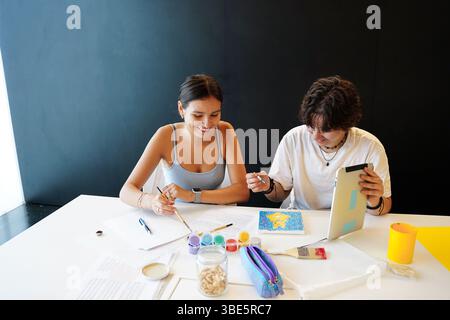 Two teenagers, one non-binary and one female, participate in artistic activities at school. They are focused on painting and using various study mater Stock Photo