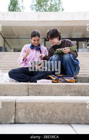 Two cheerful teenagers, a non-binary individual and a female, sit on university steps immersed in their studies. They are engaged in a discussion with Stock Photo