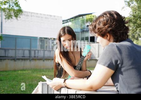 A female plays a ukulele while a non-binary teenager with short hair looks on intently, holding papers, both seated outdoors with a school building in Stock Photo