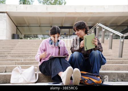 Two teenagers, a non-binary genderqueer individual and a female, are focused and engaged in studying together with notebook and digital tablet on the Stock Photo