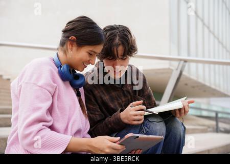 Two teenagers, a non-binary individual and a female, are studying together outdoors, using a tablet and notebook. The environment is casual, suggestin Stock Photo