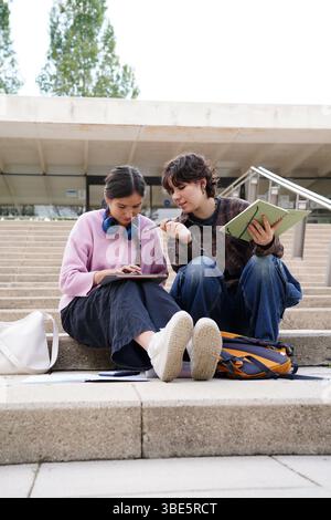 Two teenagers, a non-binary individual and a female, sit on school steps deeply engaged in study with a tablet scree, discussing educational content w Stock Photo