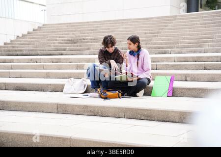 Two teenagers, a non-binary individual and a female, are engaged in a collaborative study session on university steps, surrounded by a tablet, noteboo Stock Photo