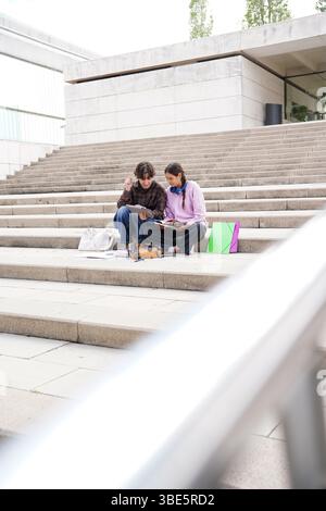 A non-binary teenager with short hair and a female teenager sit closely on university steps, engrossed in studying from notes and a tablet, depicting Stock Photo