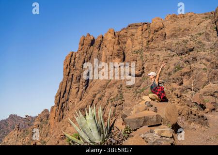 Side view of excited female hiker admiring the rugged terrain of Pico de las Nieves, Gran Canaria. She sits on a rocky landscape near agave plants, po Stock Photo
