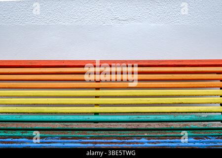 Vibrant Rainbow Colored Wooden Bench Against A Textured White Wall. Bright Colors, Abstract Pattern, Public Seating, Urban Detail Stock Photo