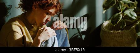 Person with red hair sitting by plant, contemplating something in sunlight. Sunlight casting shadows against the wall, creating an introspective atmos Stock Photo